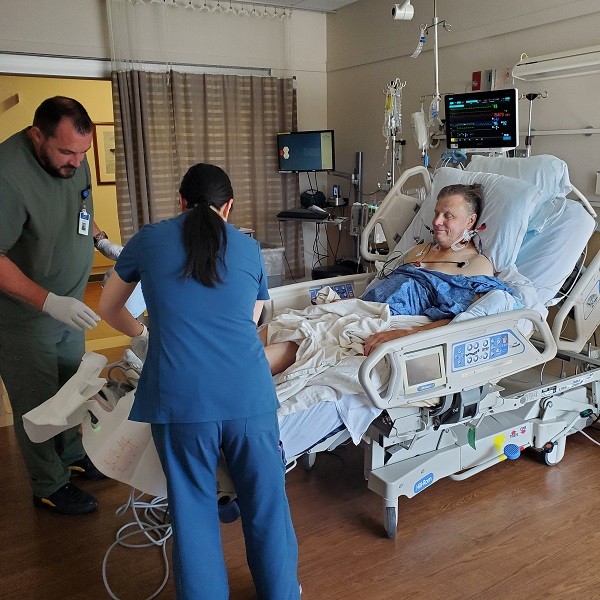 Two healthcare workers attend to a smiling patient in a hospital bed connected to medical monitors