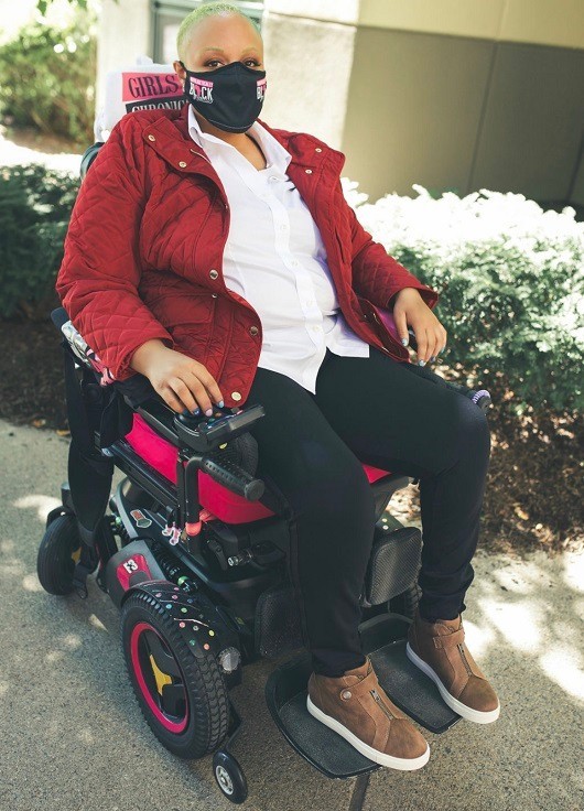 Keisha Greaves sits in her black power wheelchair wearing brown suede zip-up sneakers, blank pants, a white collared shirt, and a red quilted jacket. She has light brown skin, painted fingernails, brown eyes, and very short curly green and blue dyed hair. She wears a black mask across her nose and mouth from her Girls Chronically Rock fashion collection.