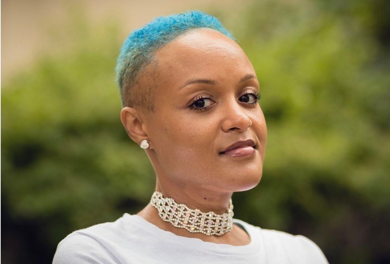 In a horizontal portrait, Keisha Greaves looks into the camera with dark brown eyes, light brown skin, short curly blue hair, and sparkling jewelry at her ears and throat.