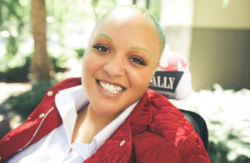 In a soft-focus horizontal portrait, Keisha Greaves grins in a white collared shirt and red quilted jacket. She has light brown skin, dark brown eyes, a stud in her nose, and green dyed short curly hair.