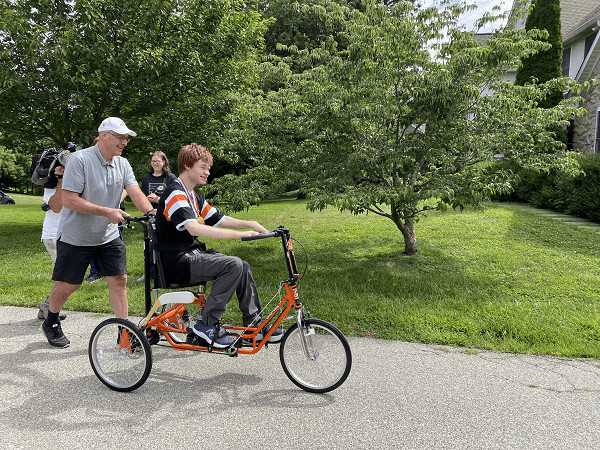 Flyers Alumni Brad Marsh pushes 18-year-old Patrick on his new adaptive bike outside of his home. Brad wears a gray polo shirt, black shorts, back sneakers, and a white cap and his glasses. Patrick wears a black and orange Flyers jersey. His bike is bright orange with black accents. Both are smiling.