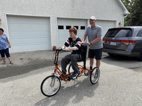 Flyers Alumni Brad Marsh pushes 18-year-old Patrick on his new adaptive bike outside of his home. Brad wears a gray polo shirt, black shorts, back sneakers, and a white cap and his glasses. Patrick wears a black and orange Flyers jersey. His bike is bright orange with black accents.