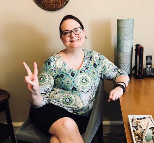 Linda Jara gives a peace sign wearing a colorful printed top and black skirt and leaning on a brown desk. She has light skin, brown hair, and blue-gray eyes behind black rimmed glasses.