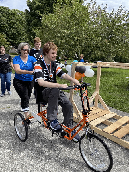 18-year-old Patrick grins as he rides his orange and black Freedom Concepts adaptive bike. He’s wearing a black and orange Flyers jersey and a replica gold medal. He’s being pushed by a loved one with gray hair and a blue shirt. Behind her are his brother and sister, each wearing matching Flyers jerseys. Brad Marsh is walking up his driveway in the far distance.