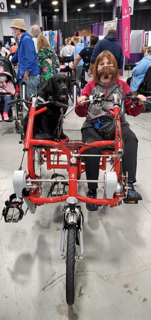 Help Hope Live client Amy Sherwood and her service dog Dolly Pawton on a red bicycle style mobility device at the 2022 Abilities Expo New York Metro