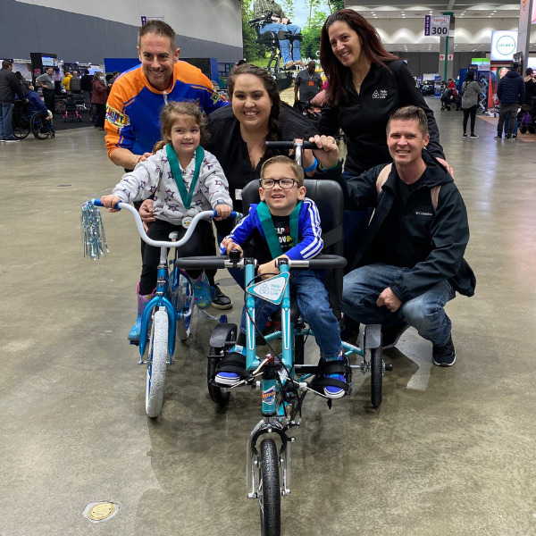 4-year-old Austin sits on his new teal adaptive bike and smiles with a medal around his neck with his twin sister on her own traditional bike, family members, and Help Hope Live Executive Director Kelly Green on the expo floor at Abilities Expo LA.