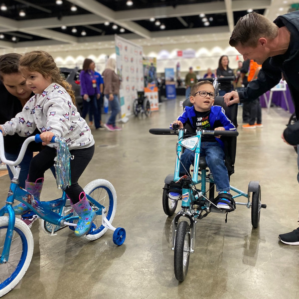 4-year-old Austin smiles atop his new teal adaptive bike as family members help him and his sister to ride—she sits on a traditional bike of her own with training wheels.