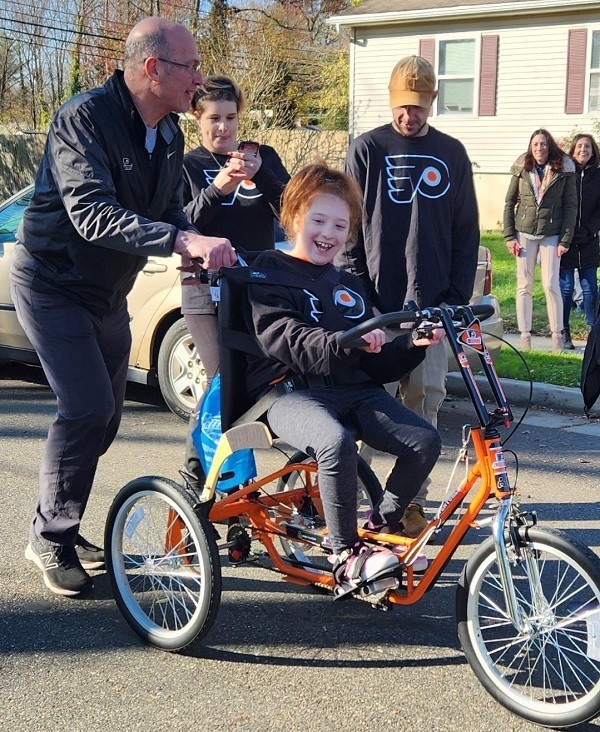 Flyers Alumni Brad Marsh pushes 9-year-old Raelynn as family members and neighbors look on. Raelynn is smiling from ear to ear as she sits atop the bright orange ad black adaptive bike.