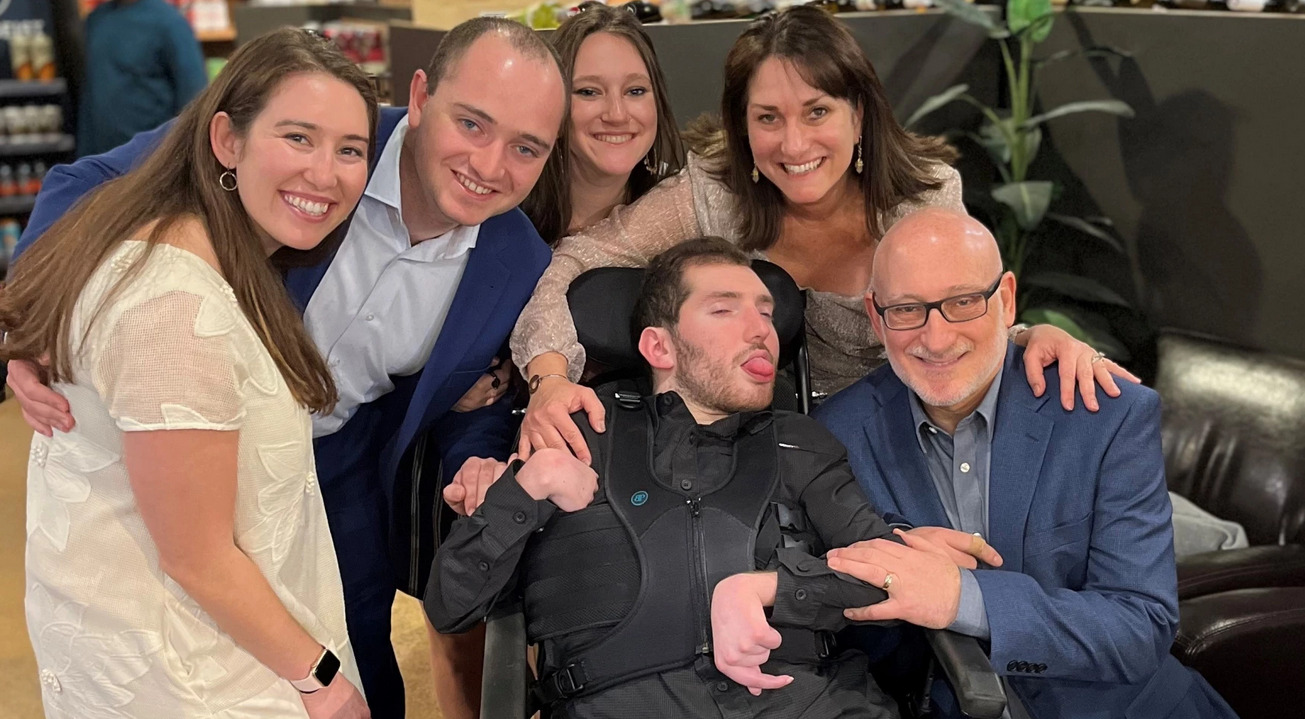 22-year-old Ethan Kadish is strapped int a black harness and is seated in a black wheelchair. He has short curly brown hair, light skin, faint stubble, and a pink tongue protruding from between his lips. Surrounding him are his parents and other family members, dressed in formal attire and smiling.