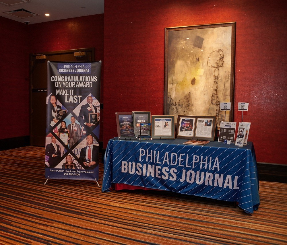 A welcome table in a hotel/casino lobby has a Philadelphia Business Journal tablecloth with a table covered in framed awards and magazine excerpts. A standing banner reads Congratulations on Your Award – Make It Last with photos of prior-year recipients in formal attire holding their awards.
