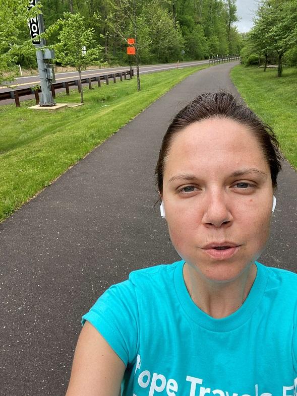 A woman with dark hair pulled back and white airpods frowns at the camera outdoors. She is wearing a teal t-shirt.