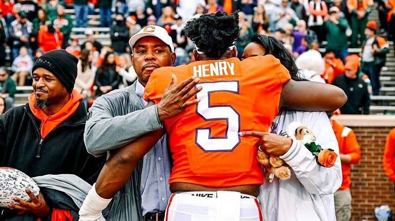 Former assistant football coach and kidney transplant recipient Keith Henry has a look of intense pride on his face as he hugs and pats the back of his son, KJ, who is wearing a bright orange Clemson college football jersey with the number 5. KJ's mother is on his other side hugging him. Keith, KJ, and his mother all have brown skin and brown hair.