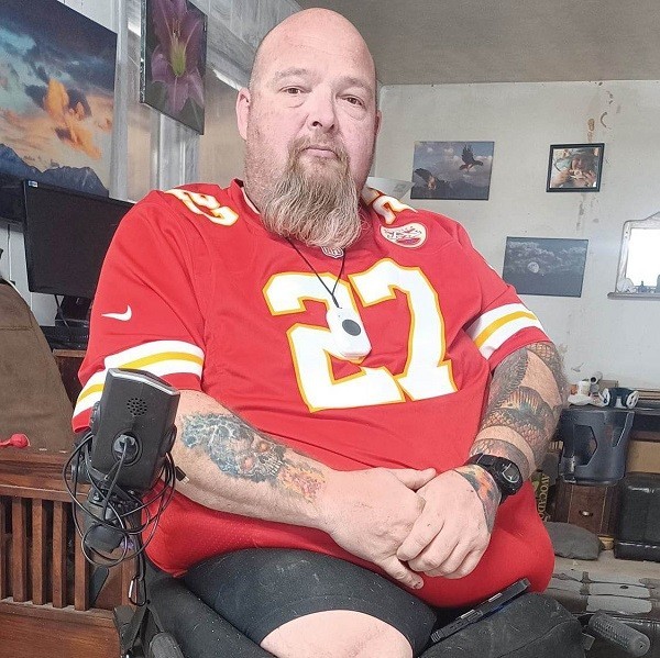Double amputee Michael Bechaver is seated in a black power chair. He has light skin, a reddish-brown goatee, a bald head, and tattoos on his arms. He wears a red sports jersey with the number 27 on the front. He has one leg amputated at the hip and one partially amputated leg that extends halfway between hip and knee.