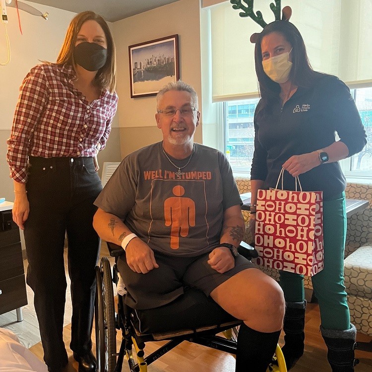 Help Hope Live's Shannon Shensky and Kelly Green deliver a Bags of Hope gift to an individual at Magee Rehabilitation. He has light skin, gray hair, and a short gray beard and glasses and is seated in a black wheelchair. His shirt reads Well I'm Stumped - a play on the fact that the man has one amputated leg.