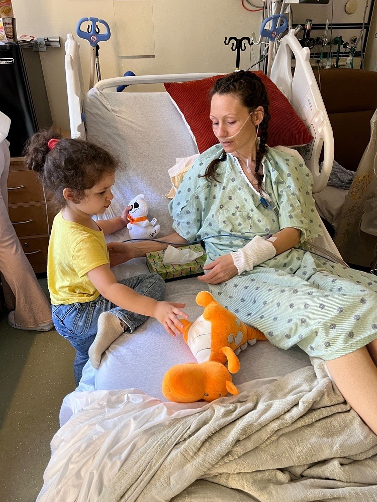 Pamela has light skin, curly dark brown hair in braids, and a hospital gown as she sits in her hospital bed and plays with toys with her young daughter, who also has light skin and curly dark brown hair.