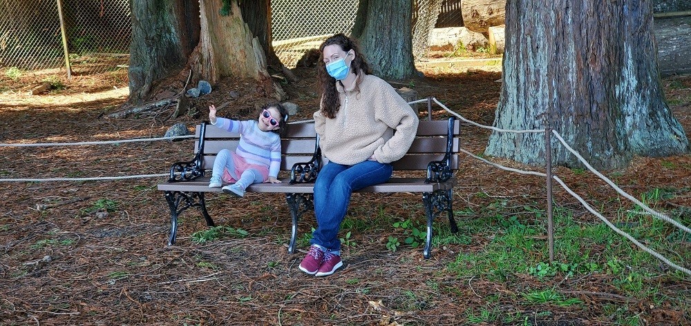 Pamela wears jeans, sneakers, and a tan sweater and sits on a park bench in a wooded area beside her youngest daughter, who wears pastel stripes and children's sunglasses. Pamela and her daughter both have curly brown hair and light skin.
