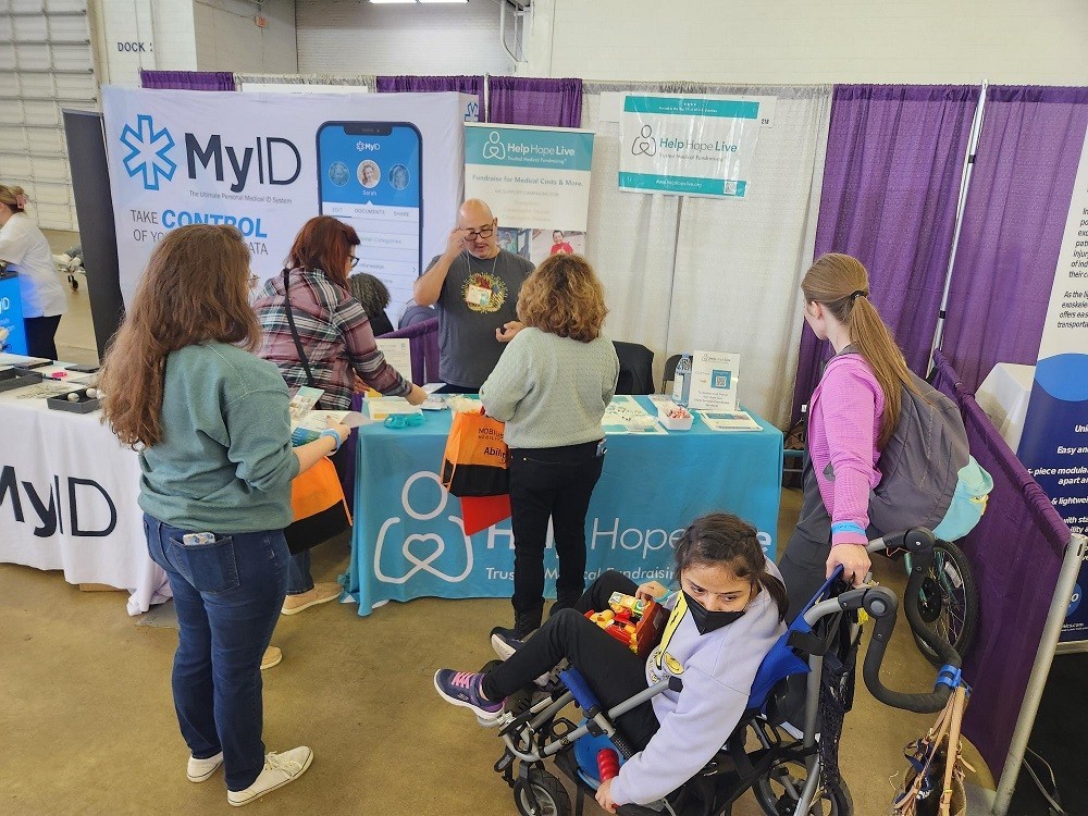 Kevin Lopez is staffing the Help Hope Live booth at Abilities Expo Dallas. Several people are in front of the booth, including four female-presenting attendees who are standing and one child in a stroller wearing a black face mask. The Help Hope Live booth includes signage, bracelets, and brochures. Kevin has light brown skin, black rimmed glasses, and light gray facial hair.