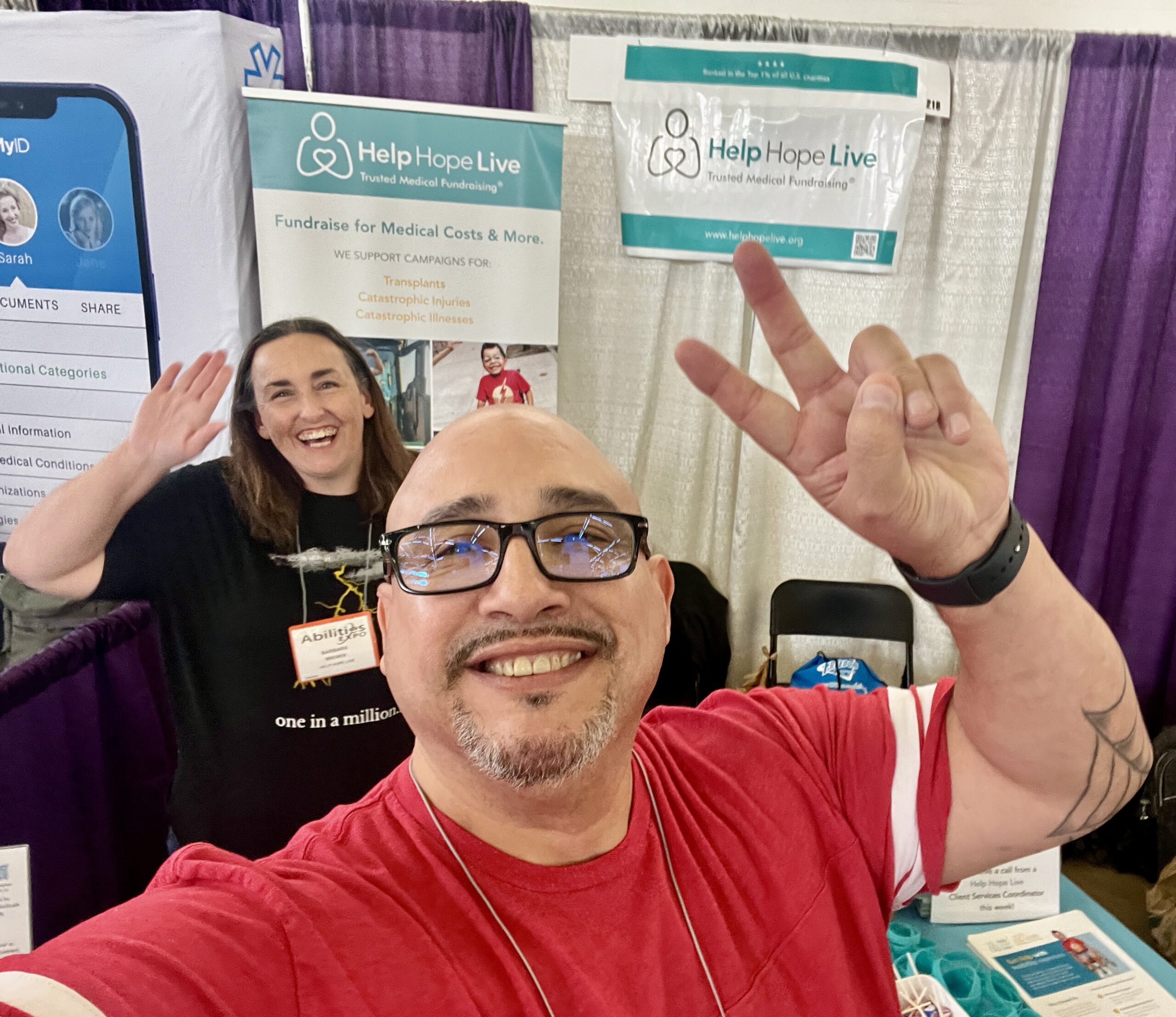 In a selfie at Abilities Expo Dallas in front of the Help Hope Live booth banners, Kevin Lopez and Barbara Brewer smile. Kevin has light brown skin, a bald head, and a short gray goatee with a bright red shirt, and he’s giving a peace sign. Behind him Barbara has light skin and shoulder-length brown hair with an abilities Expo badge—she is waving.