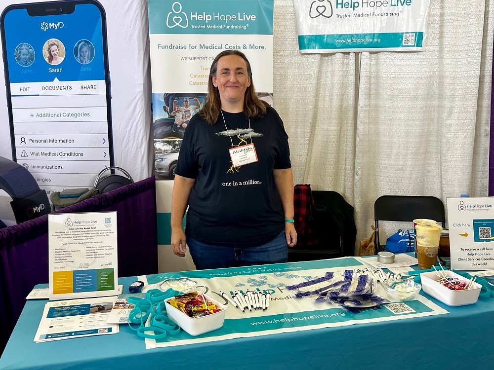 Barbara Brewer is at the Help Hope Live table and booth at Abilities Expo Dallas 2023. She has light skin, shoulder-length brown hair, light eyes, jeans, and a black t-shirt that has a lightning graphic and text