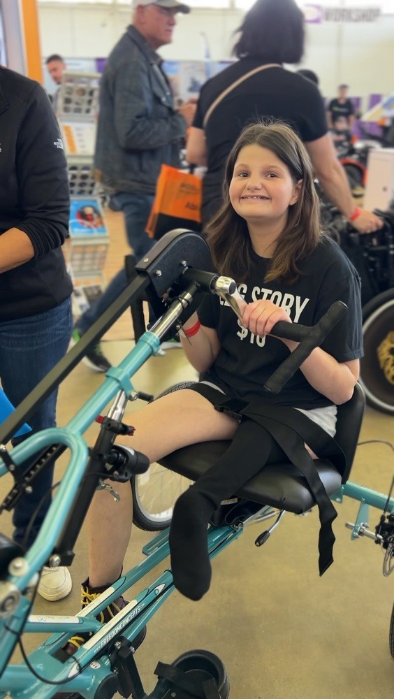 15-year-old Hallie is grinning as she rides her new teal Help Hope Live-themed handcycle adaptive bike at Abilities Expo Dallas. Hallie has light skin, a big smile with braces, and wavy shoulder length brown hair.