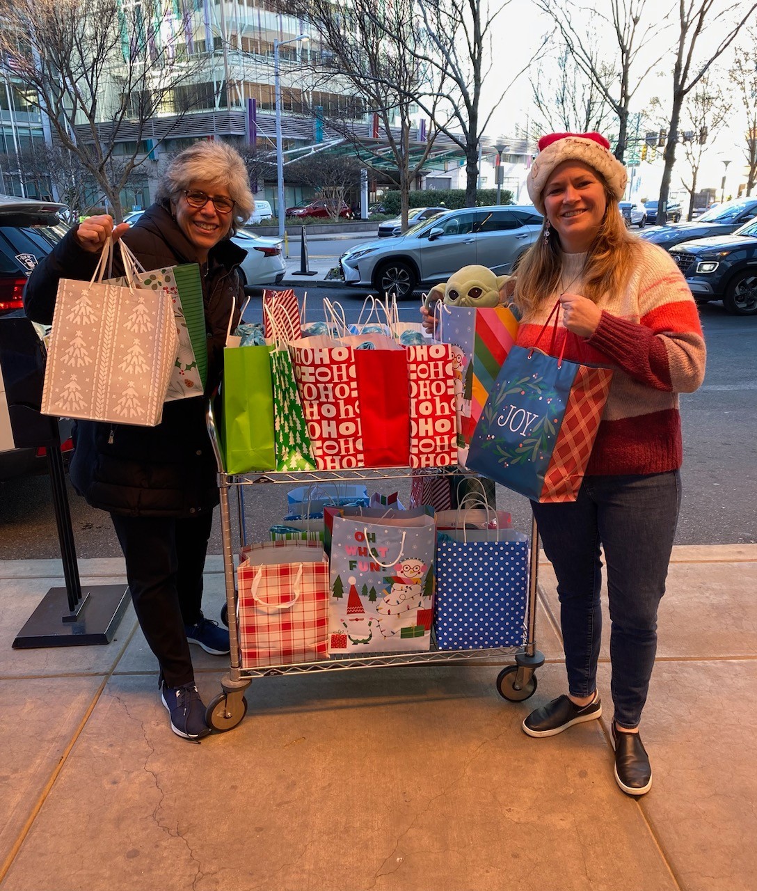 Sonny Mullen is with Deb Gordon at the Hospital of the University of Pennsylvania. On a city sidewalk, they flank a rolling cart full of brightly colored handled holiday gift bags. Deb has light skin, short gray hair, and glasses. Sonny has light skin, blonde hair past her shoulders, and a Santa hat. They wear warm winter casual clothes.