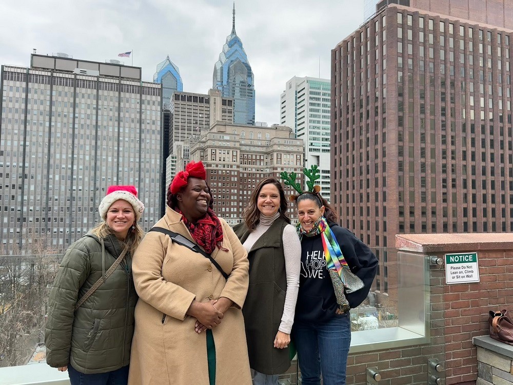 Four Help Hope Live staff members are pictured on the rooftop of Magee Rehabilitation with a portion of the Philadelphia city skyline behind them in winter. Sonny has light skin, blonde hair, and a Santa hat. Vanessa has dark skin, a big red bow, and a camel coat. Shannon has light skin, highlighted shoulder length brown hair, and a cream turtleneck with a vest. Kelly has light skin, dark hair pulled back, a Help Hope Live black hoodie and jeans, a rainbow scarf, and a green reindeer antler headband.