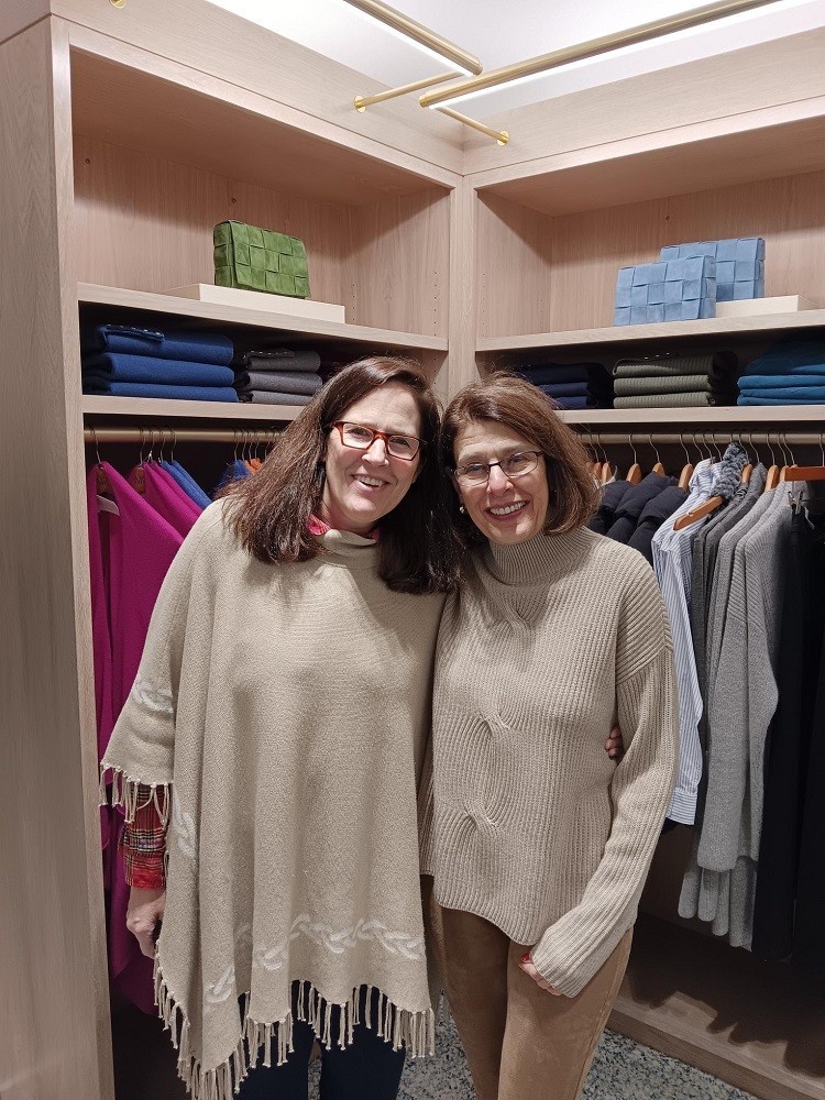 Help Hope Live founding supporter Christine Kanter is with the J.McLaughlin store manager standing in a carpeted shopping area with sweaters and formal shirts on hangers behind them. The store manager wears a a cream tassled poncho and has light skin, tortoiseshell glasses, dark eyes, and brown hair to her shoulders. Chris wears brown suede pants and a cream turtleneck with light skin, brown hair ending just before her shoulders, dark eyes, glasses, and red lipstick.