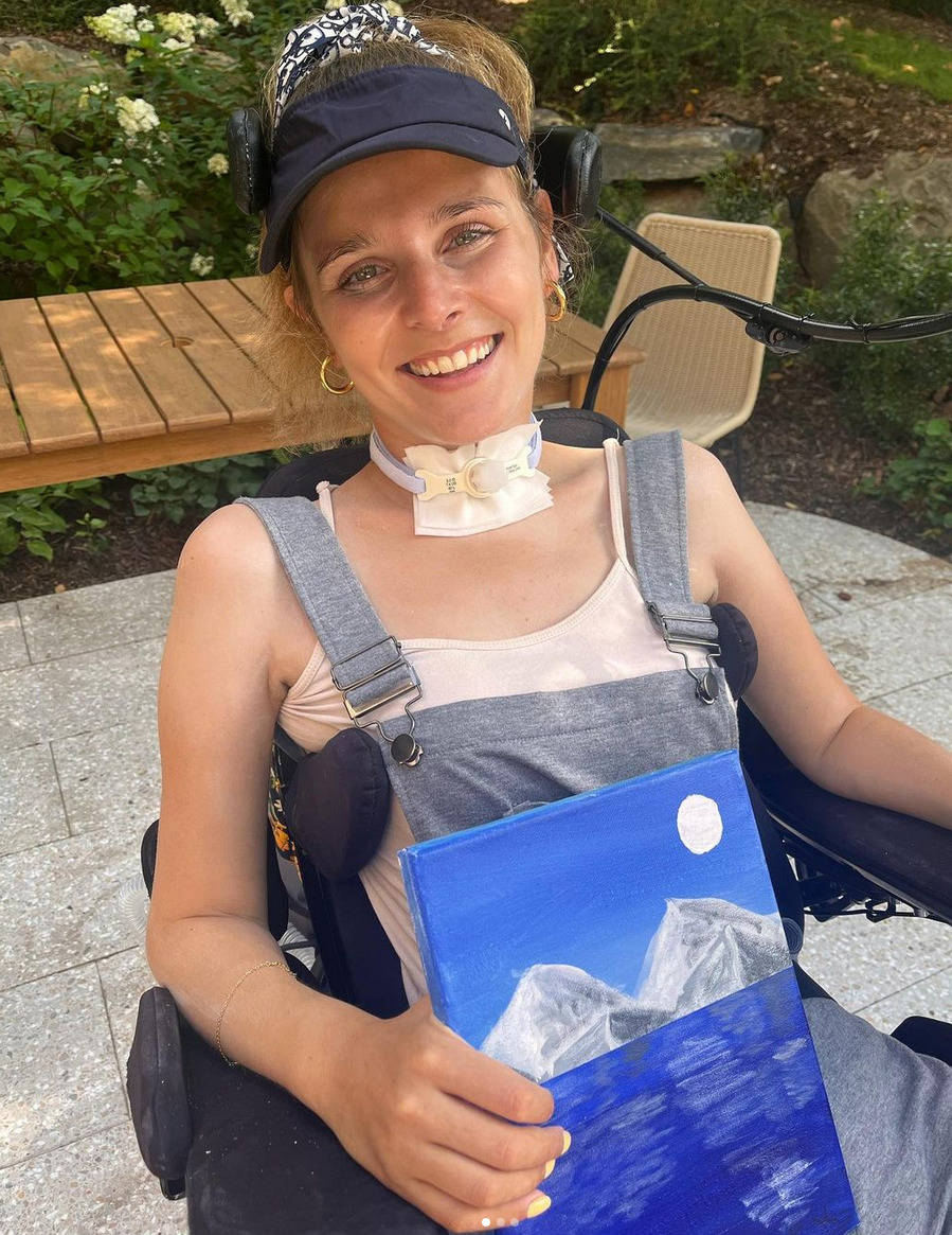 Rebecca smiles as she holds one of her mouth-painted pieces in her right hand. The piece is a scene of mountains reflecting into water under the light of the moon in shades of white, silver, deep blue, and light blue. Seated in her black power chair with a mouth stick for mobility and a patch at her neck for a trach, Rebecca has light skin, gold hoop earrings, light eyes, a black cap, and her blonde-brown hair pulled back. She wears a sleeveless light pink blouse with gray overalls overtop.
