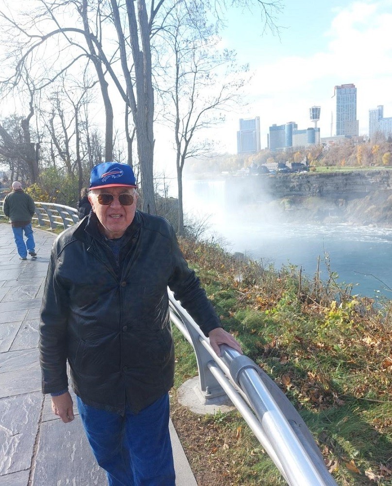 By the banks of Niagara Falls next to a handrail on a walking path, heart transplant recipient Greg Wright has light skin, short gray hair, sunglasses, and a blue baseball cap. He wears a buttoned leather jacket and blue pants.