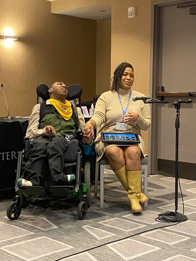 Client Noah Williams and his mom are presenting at Reeve Summit 2024. Noah has brown skin and a bright yellow bandana over a forest green vest. He is seated in a black wheelchair gazing upward. His feet, in socks, point left and right. Mom Naomi has brown skin, a cream colored turtleneck dress, yellow cowboy boots, and a speech communication device on her lap. She grasps Noah's hand.