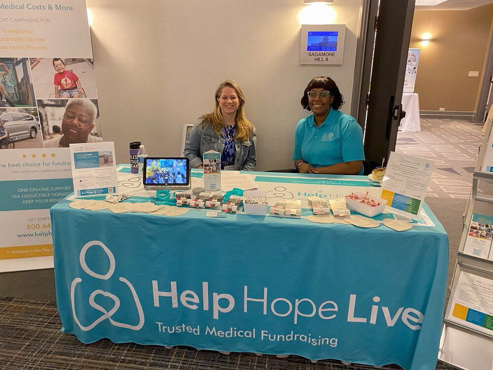 At the Help Hope Live booth of the Reeve Summit 2024, which is branded with teal and the tagline Trusted Medical Fundraising and a table covered in brochures, pens, and other swag items, Sonny and client Julie Chambers are seated. Sonny has light skin, golden hair past her shoulders, a blue printed dress, and a jean jacket. Julie has brown skin, wavy shoulder-length brown hair with bangs, dazzling glasses, and hoop earrings, and she wears a teal Help Hope Live polo.