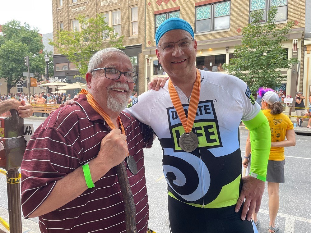 Heart transplant recipient Bill Soloway takes a photo on a suburban street with his donor's father. Bill has light skin, a blue headband, a Donate Life blue and green cycling shirt, and a medal on an orange ribbon. His donor's father has a matching medal and has light skin, glasses, short white hair, a white mustache, and a white goatee and he holds a walking stick in his right hand.