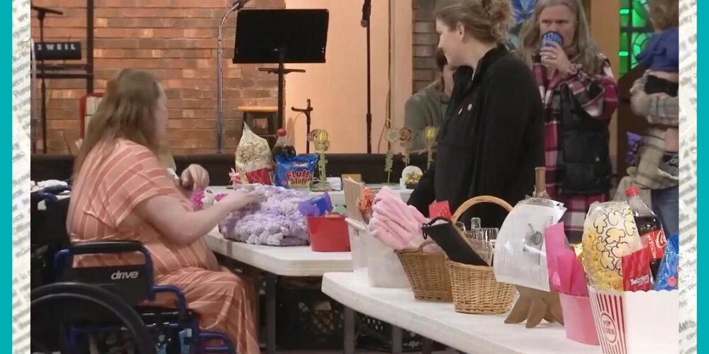 A screenshot from news coverage of a fundraiser that features raffle or auction baskets on tables and people walking around to view them and bid. Client Lisa Marie Lee is seated in a manual wheelchair with purple accents on the other side of the tables. She has light skin, wavy brown hair past her shoulders, and a striped salmon pink dress.