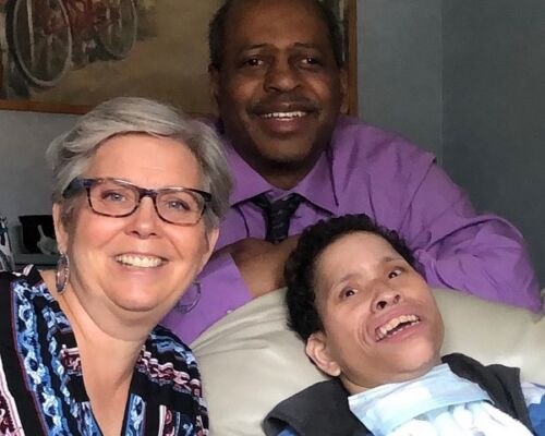 Three adults smile at the camera; one is reclining with a medical tube visible