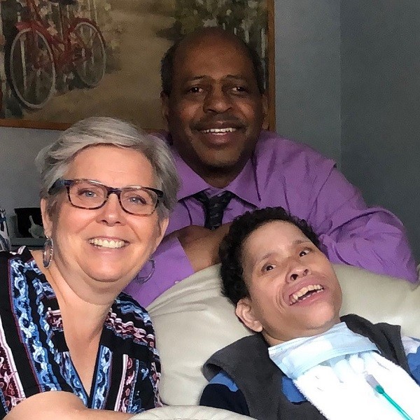 Three adults smile at the camera; one is reclining with a medical tube visible