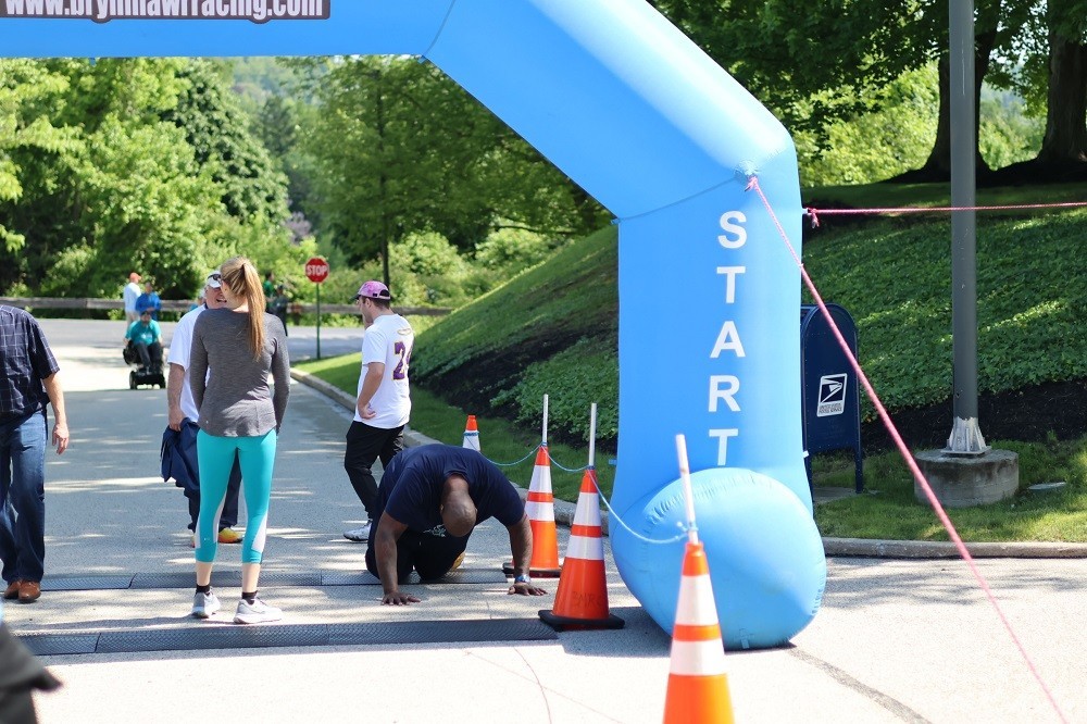 In a funny photo, Barret Brooks falls to his hands and knees in relief as he reaches the finish line of the Hope Travels 5K event in 2024. Surrounded by other participants in athletic gear of various ages, Barrett has brown skin and wears a navy t-shirt.