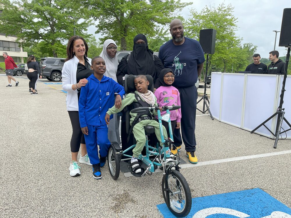Outdoors at Hope Travels 2024, Help Hope Live Executive Director Kelly L Green and Barrett Brooks pose with 8-year-old Khuwaylid and his family. Khuwaylid is seated on his new customized teal adaptive bike and has brown skin and a pistachio green sweatsuit. His mother wears a black niqab and has brown skin. The women to her right wears a gray hijab and has brown skin and clear frame glasses. His twin brother has brown skin and a bright blue two-piece jumpsuit. His younger sister wears a black hijab and a bright pink sweatshirt and glasses. Barrett has brown skin and a short black and gray goatee. Kelly has light skin, brown hair past her shoulders, and a black Help Hope Live polo.