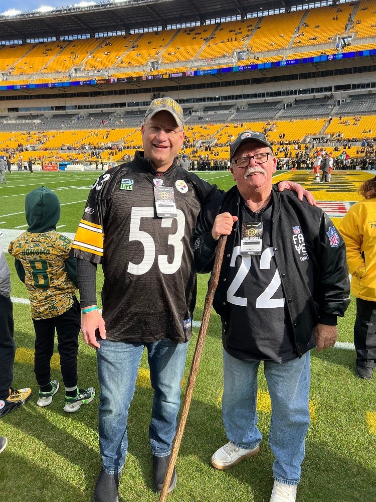 Heart transplant recipient Bill Soloway is on a Pittsburgh Steelers football field with his heart donor's father. Bill has light skin and wears jeans and a number 53 Steelers jersey. His donor's father has light skin, a white mustache, glasses, and a number 22 Steelers jersey.