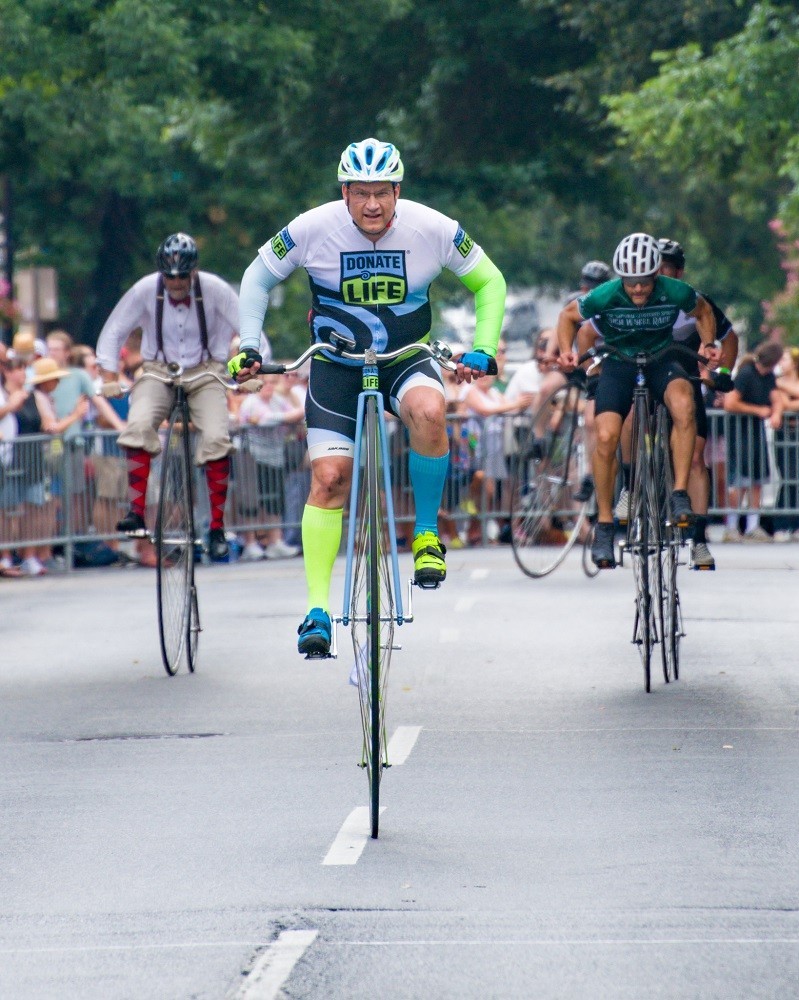Heart transplant recipient Bill Soloway leads the way during a street race for high wheel bicycles. He has light skin, and Donate Life themed cycling gear in white, blue, and green.