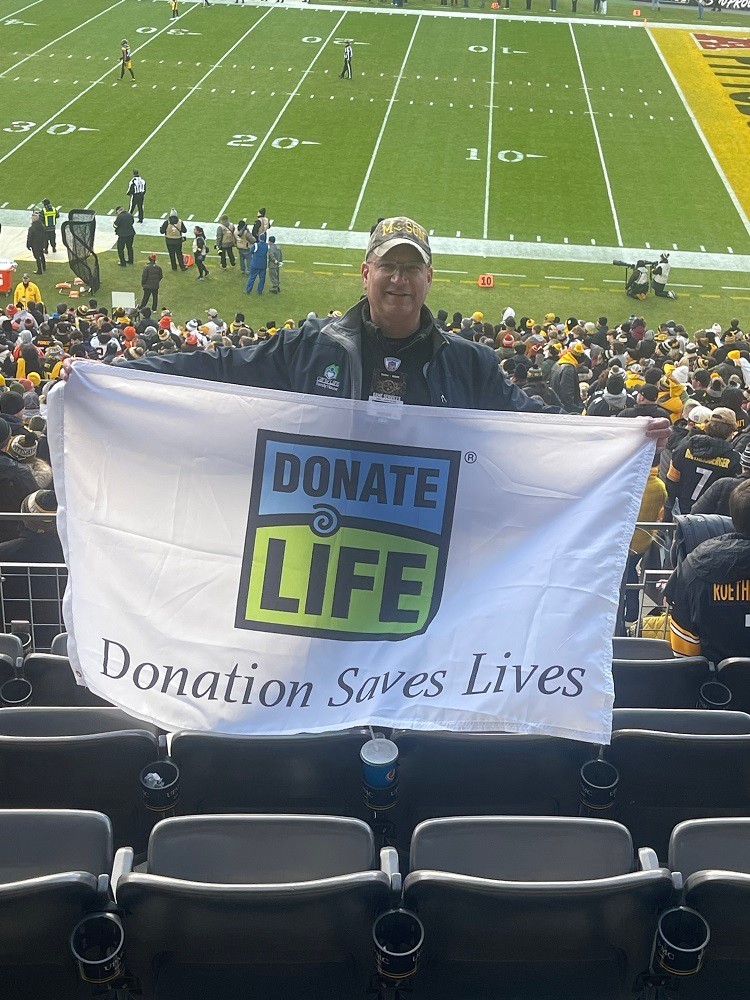 In the stands of a football event, Bill Soloway holds a large flag that reads Donate Life: Donation Saves Lives. The heart transplant recipient has light skin, glasses, and a ball cap.