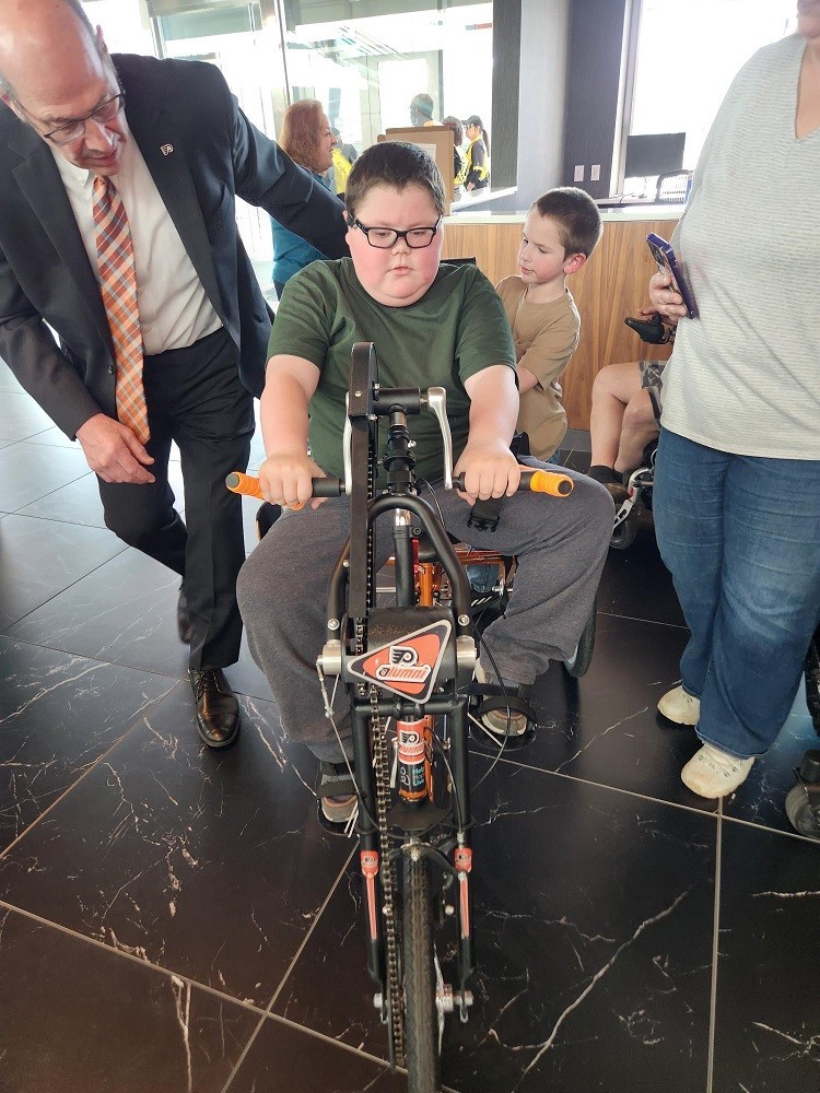 Flyers Alumni Brad Marsh gives Dylan McDonnell a push on his new Flyers themed adaptive bike. Brad has light skin, glasses, gray hair, and a suit. Dylan is a teen with light skin and glasses and short brown hair.