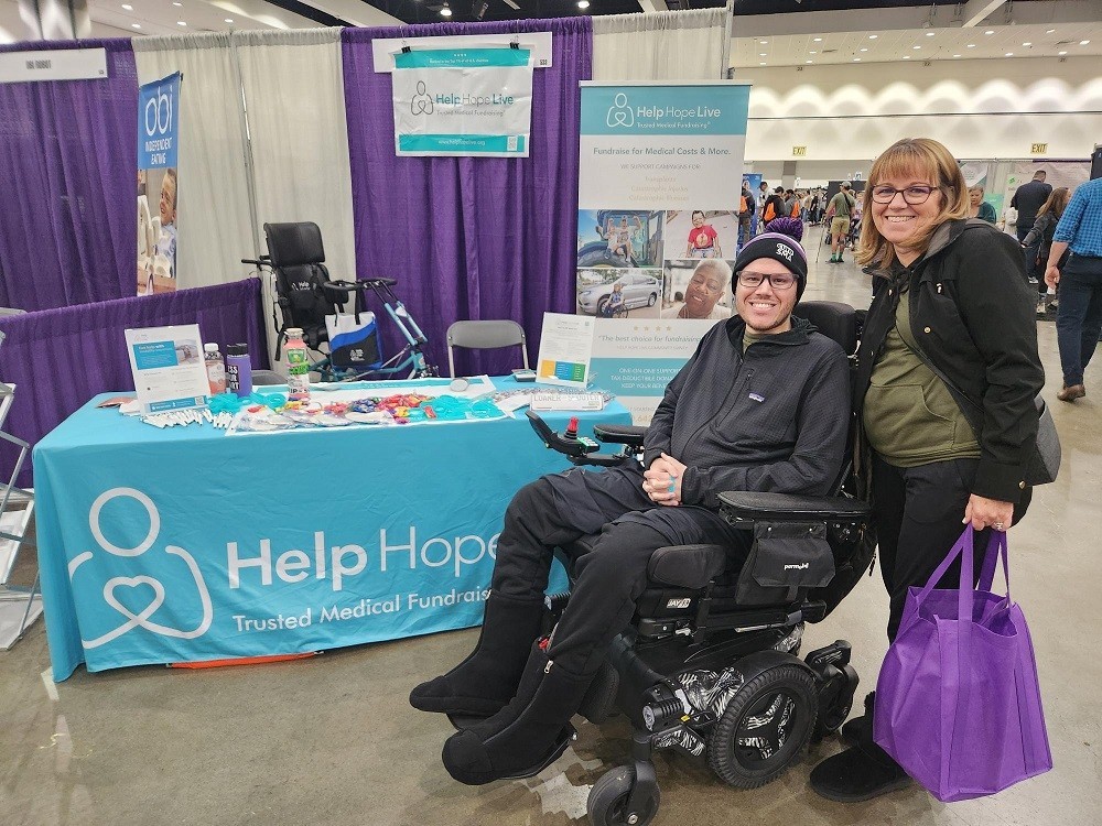 At the Help Hope Live booth at Abilities Expo LA 2024, which is branded with Help Hope Live teal and the tagline Trusted Medical Fundraising, client John Ekhart is with his mother seated in his black power chair. John has light skin, glasses, and a beanie with black warm booties. Standing beside him, his mom has light skin, shoulder-length light red hair, and glasses.