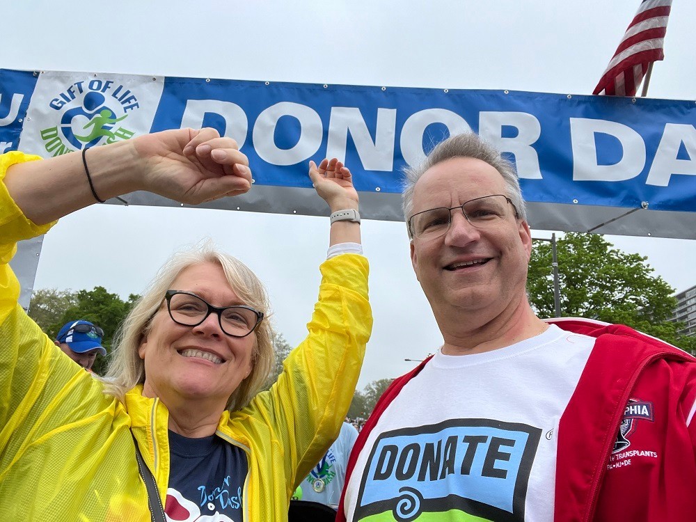 heart transplant recipient Bill Soloway and partner Kathy take a selfie at the Donor Dash. Bill has light skin, short gray hair, glasses, and a Donate Life t-shirt. Kathy has light skin, shoulder-length blonde-gray hair, and black-rimmed glasses.