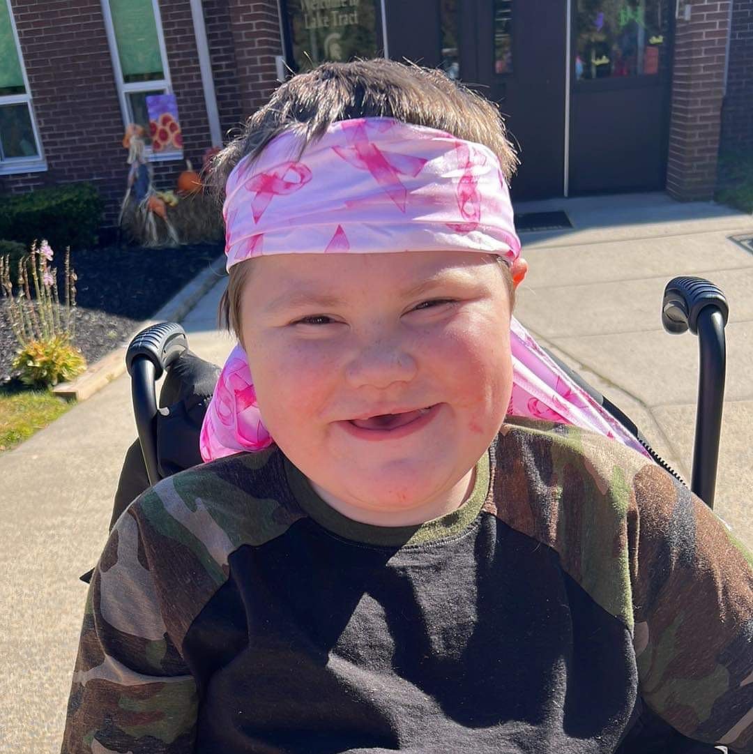A close-up portrait of Dylan McDonnell. The teen is seated in a black mobility device and has light skin, brown eyes, a big smile with some baby teeth missing, and a pink Breast Cancer Awareness themed headscarf.