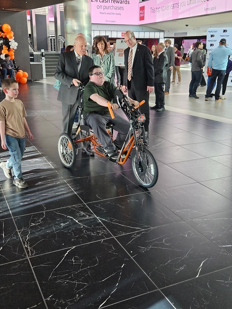 Dylan McDonnell rides his new adaptive bike at Wells Fargo center. Dylan is a teen with light skin and glasses and short brown hair. The bike is bright orange and Flyers themed with three wheels. With him are Flyers Alumni Brad Marsh and Bob Kelly.
