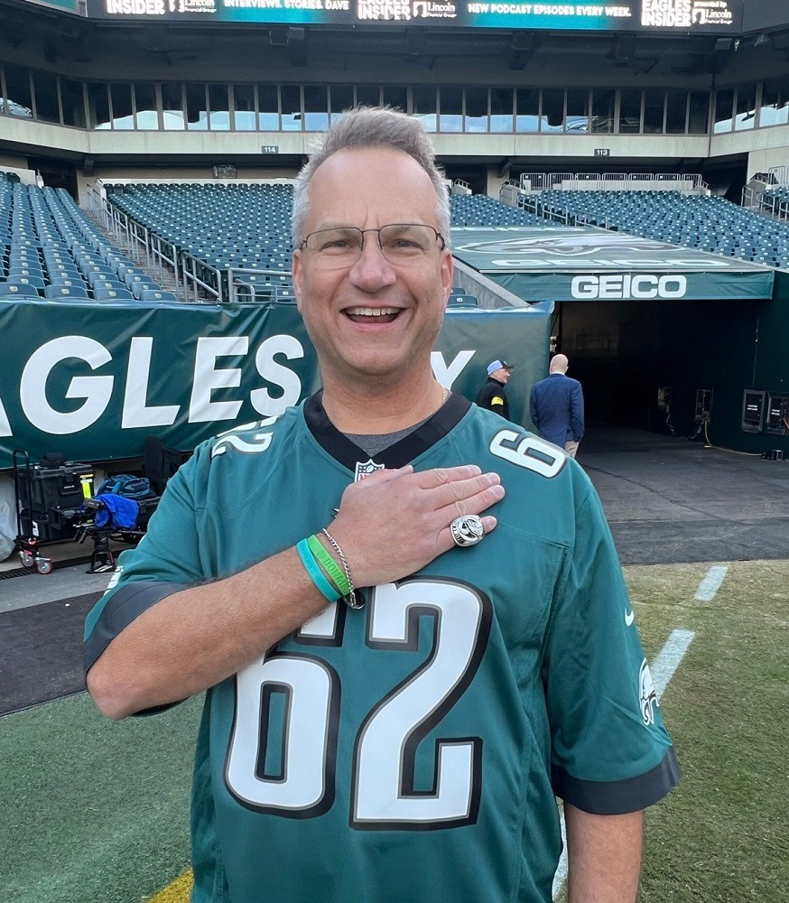 Heart transplant recipient Bill Soloway is on the Eagles football field with a number 62 Eagles jersey. Bill has light skin, short gray hair, and glasses, and he is placing his right hand over his heart to showcase his Donate Life and Help Hope Live rubber bracelets.