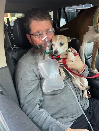 Seated in the passenger seat of a car, Edward Cardwell has light skin, glasses, and short gray-brown hair and he cradles a small dog in his left arm. He is wearing an oxygen breathing device over his nose and mouth.