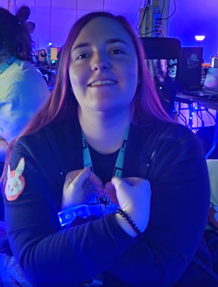 An RGB LAN 6 attendee makes a heart with her hands crossed at the wrists over her heart, replicating the Help Hope Live logo pledge figure. She has light skin, dark eyes, long pink-dyed hair, and a hoodie with the D.VA suit logo from Overwatch.