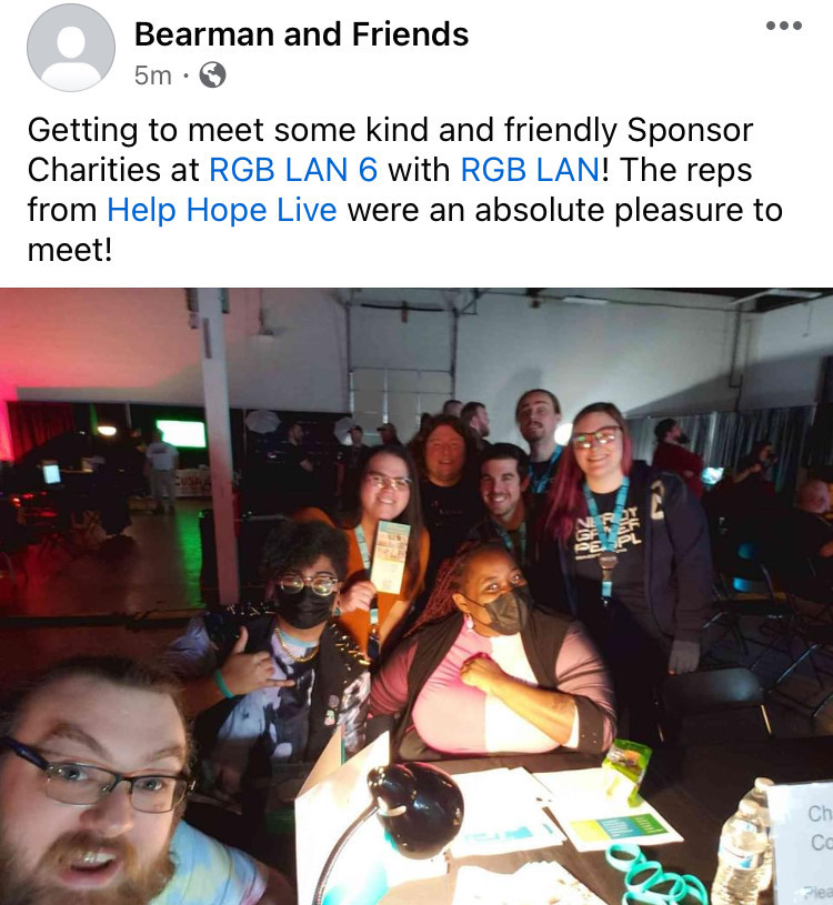 An RGB LAN 6 gaming event attendee takes a selfie in a Facebook post with Vanessa Nichols, a Help Hope Live team member, seated at a Help Hope Live table behind him surrounded by other gamers. The selfie-taker has light skin, a red-brown beard, light eyes, and glasses. Vanessa has brown skin, red dyed dreadlocks, and a color-blocked pink turtleneck dress in light and dark pink. She wears a black face mask and has brown eyes. The Facebook post is by Bearman and Friends and reads