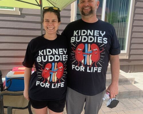 Outside on a patio, Josh Raymond is pictured with his kidney donor Shiloh. They wear matching black cartoon kidney shirts with the text Kidney Buddies for Life. Shiloh has light skin, brown eyes, and curly brown hair. Josh is a head taller than Shiloh and has light skin, blue eyes, short gray hair, and a gray beard. In the lower right corner of the photo is a logo that says Touched by Transplant with flowering green and teal vines.